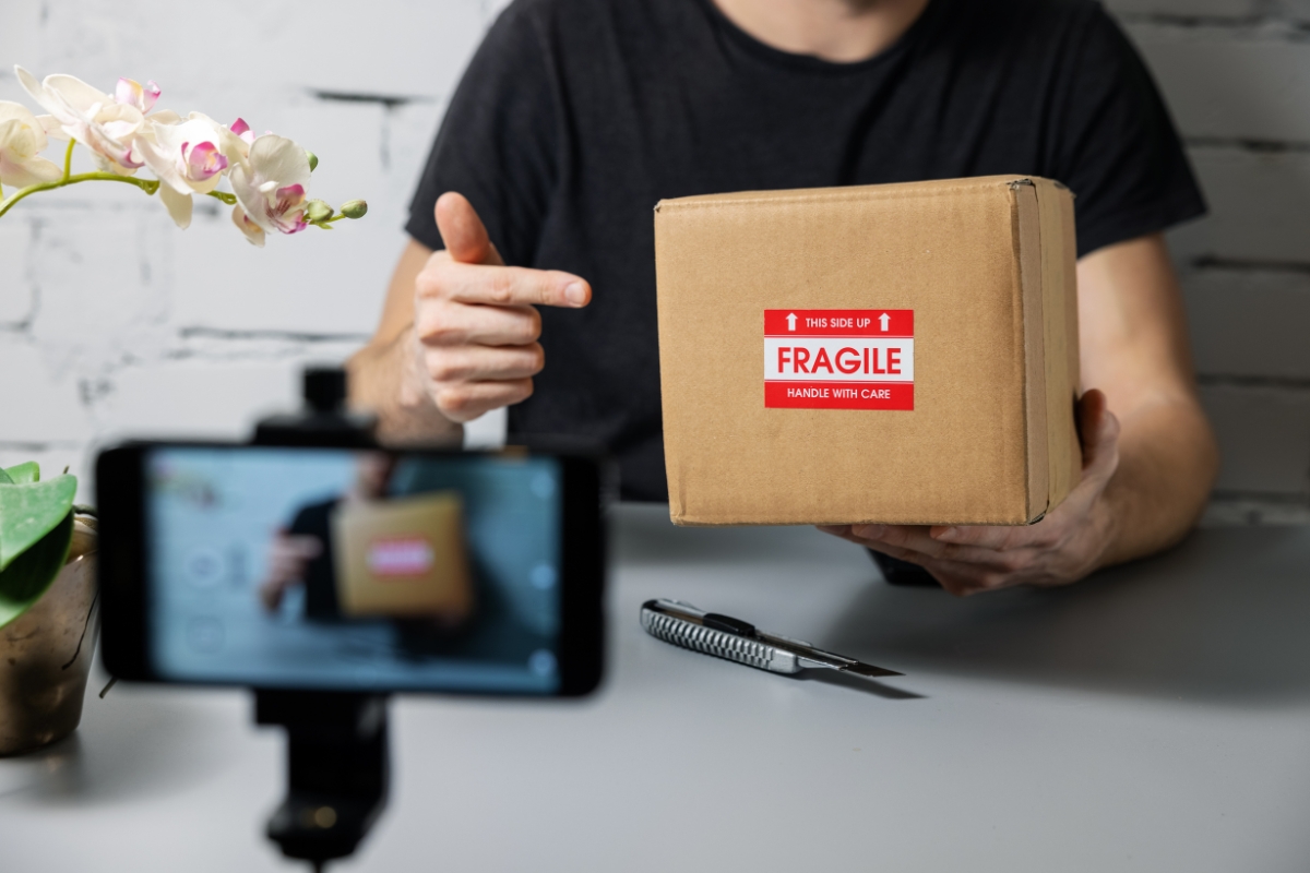 Harnessing the Power of TikTok for Business Marketing 7 A person points to a cardboard box labeled "Fragile" while recording with a smartphone on a tripod, creating Tiktok for business marketing content; a utility knife and a potted plant are on the table.