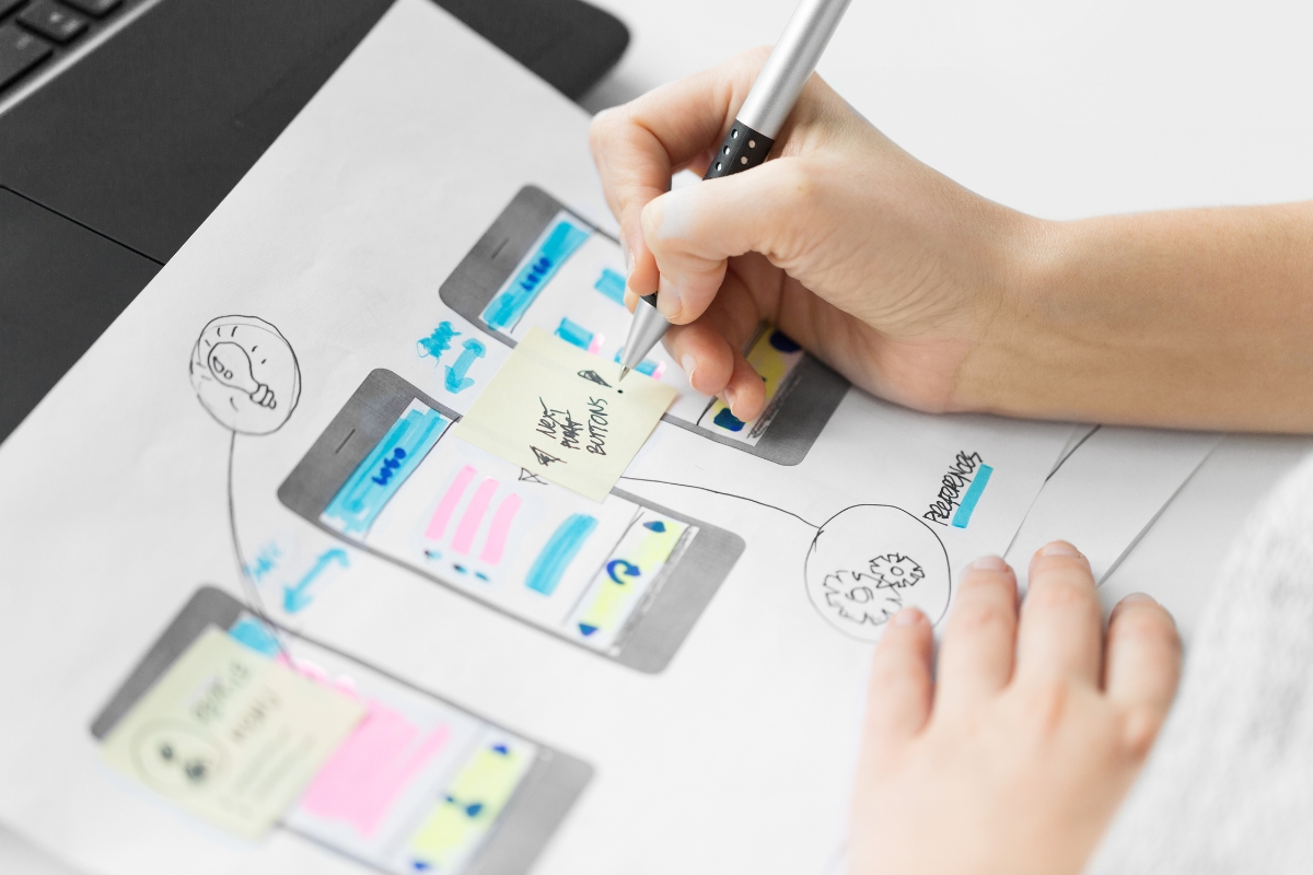 UX Conversion Rates Explained: How User Experience Drives More Sales 7 A person writes on a sticky note placed on a paper wireframe of a mobile app, highlighting the ux process to improve conversion rates next to a laptop.