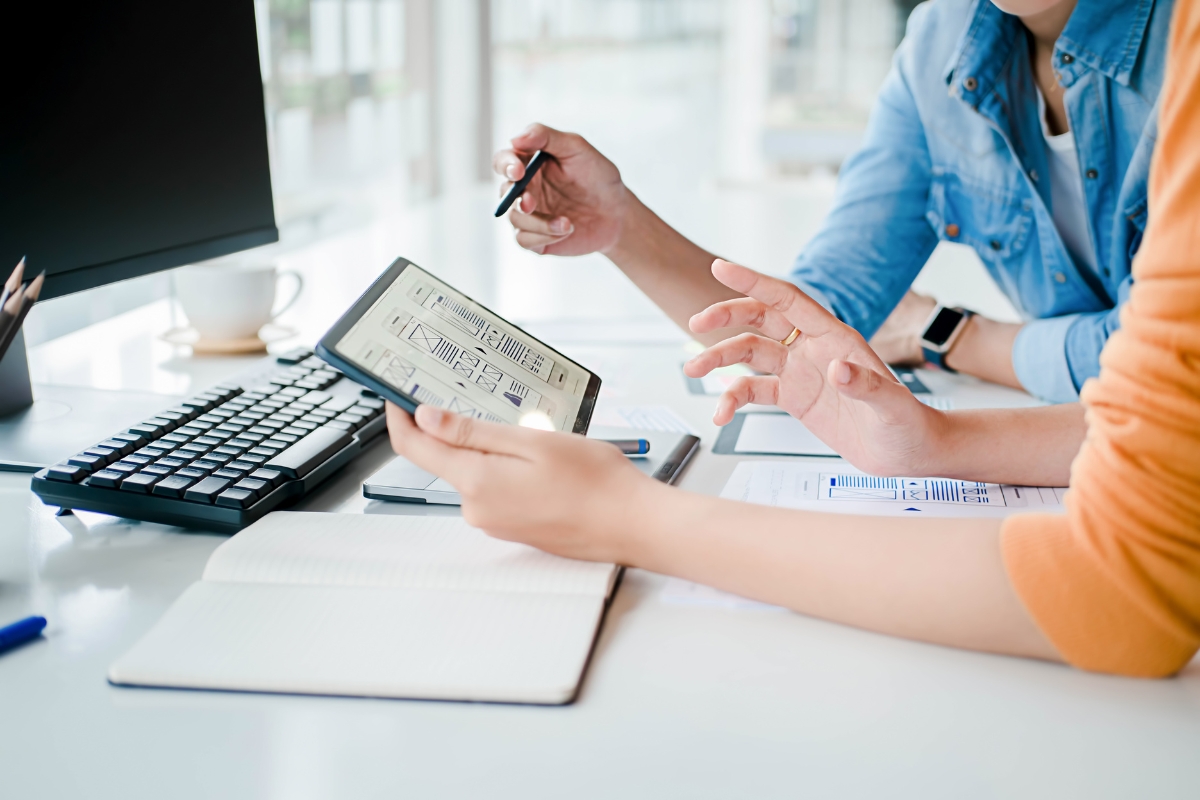 UX Conversion Rates Explained: How User Experience Drives More Sales 2 Two people at a desk review data on a tablet, with documents, a keyboard, and a notebook visible, suggesting a collaborative work discussion focused on improving UX conversion rates.