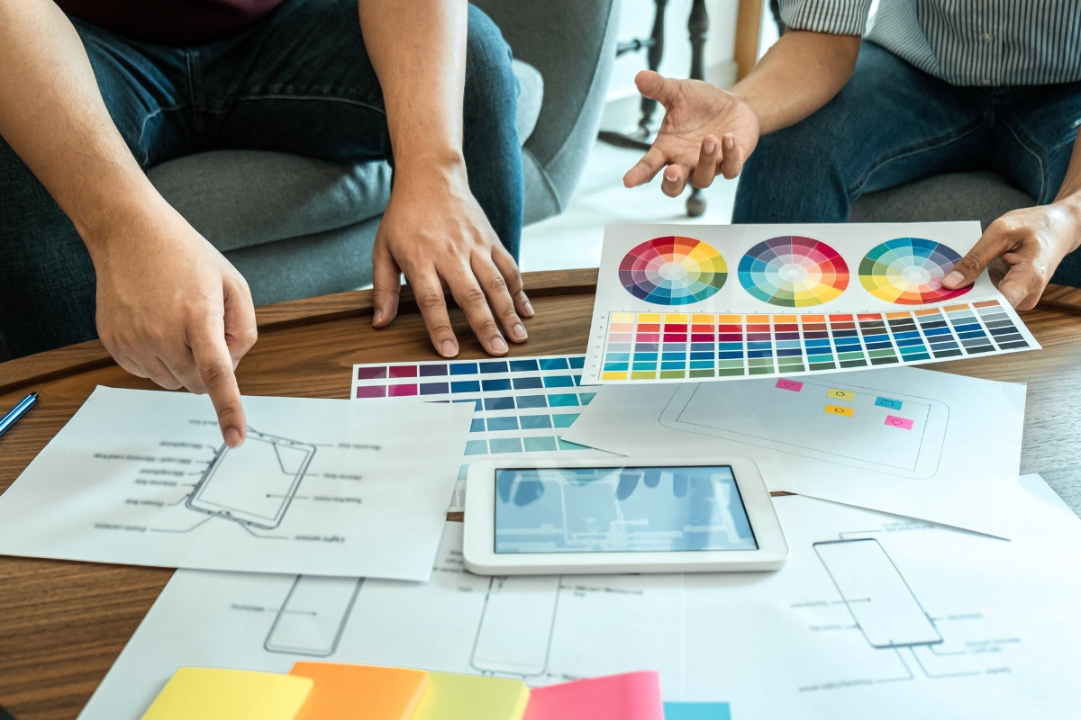 Web Accessibility: Designing for All Users 6 Two people review color swatches and wireframe sketches on a table, discussing web accessibility, with a tablet and sticky notes also visible among the design materials.
