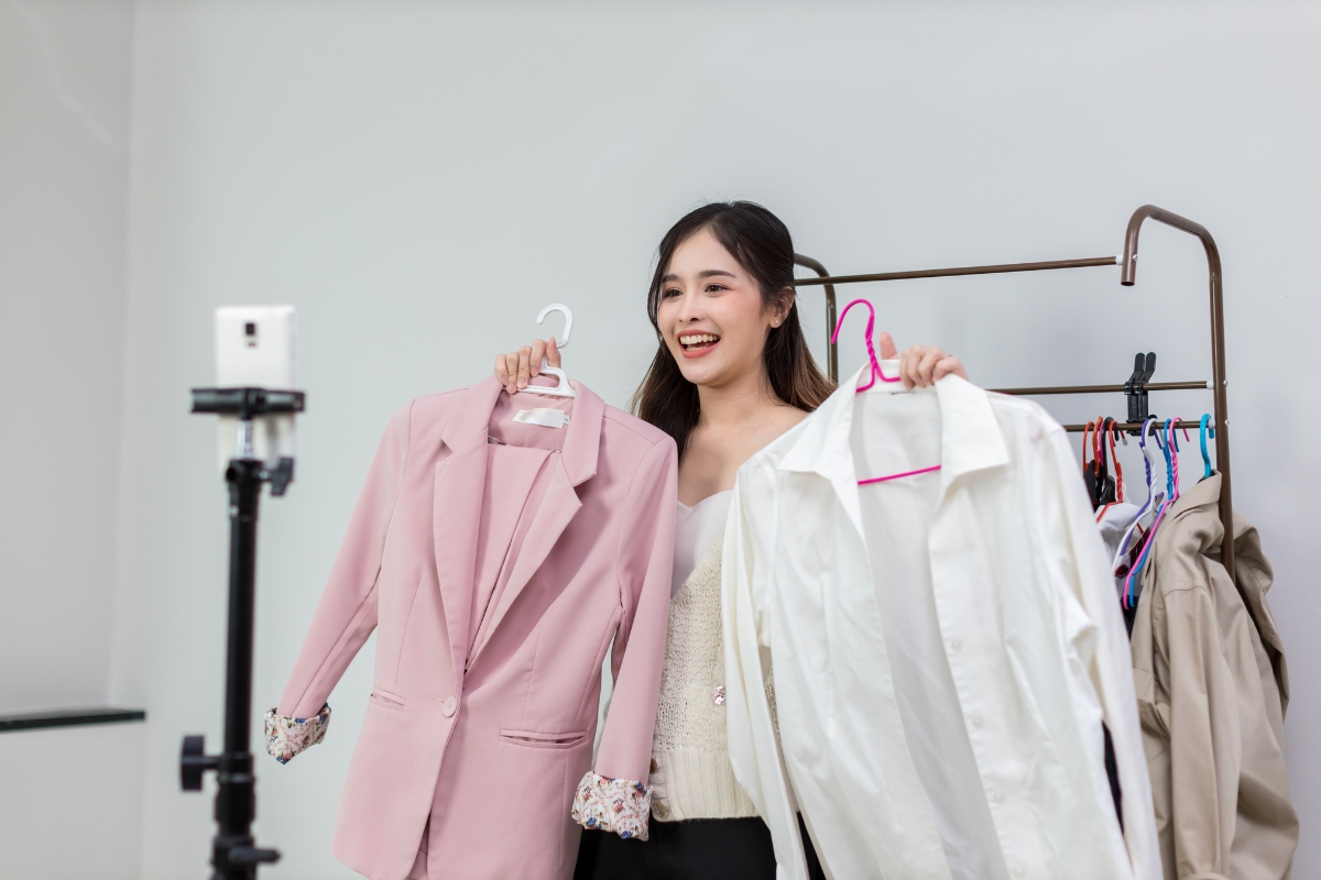 Live Streaming: Engaging Your Audience in Real-Time 4 A woman stands in front of a smartphone camera, live streaming as she holds up a pink blazer and a white shirt, with more clothes hanging on a rack behind her.