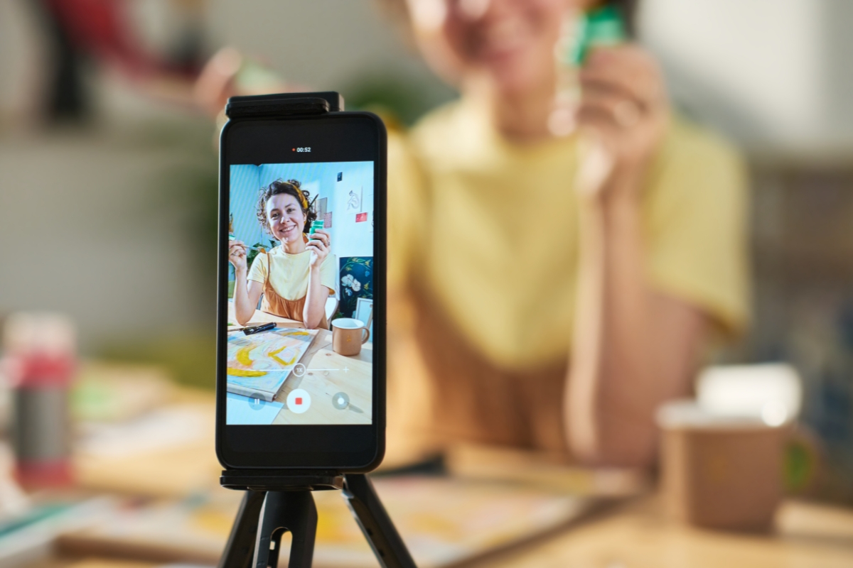 Live Streaming: Engaging Your Audience in Real-Time 5 A smartphone on a tripod captures a person in a yellow shirt live streaming at a table, holding up an object and smiling, with art supplies and a painting in the background.