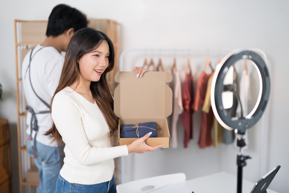 Live Streaming: Engaging Your Audience in Real-Time 8 A woman holds an open cardboard box toward a camera and ring light, livestreaming with clothing racks in the background and another person standing nearby.