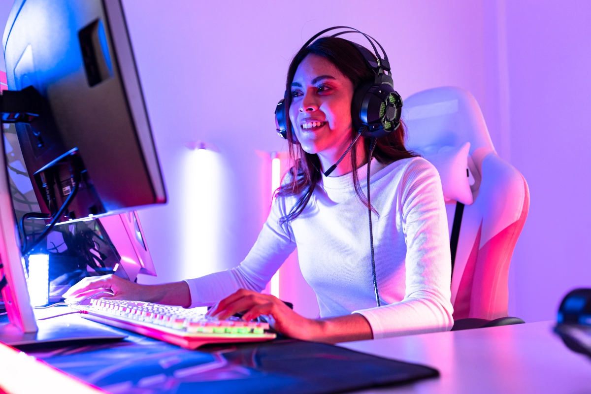Live Streaming: Engaging Your Audience in Real-Time 9 A woman wearing headphones is sitting at a desk, live streaming as she uses a keyboard and mouse while looking at a computer monitor in a room with colorful lighting.