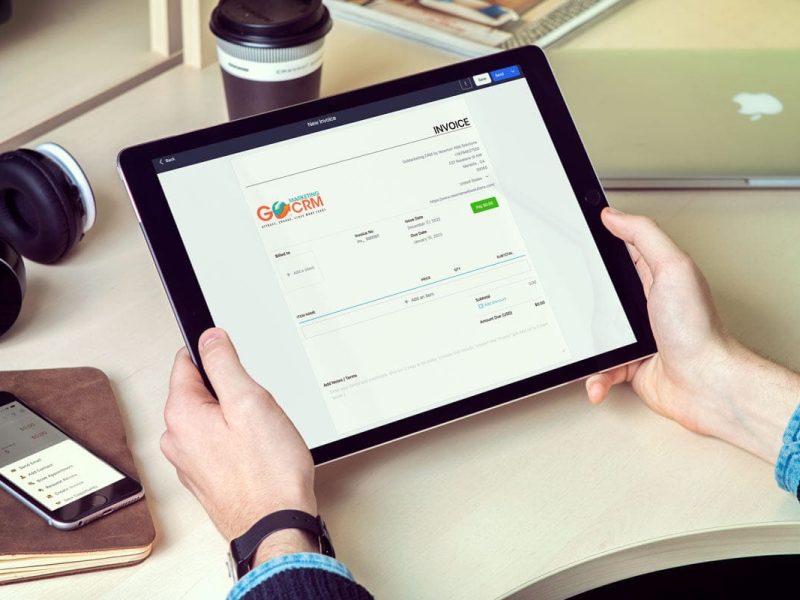 Person holding a tablet displaying an invoice. Nearby are a smartphone, headphones, coffee cup, and a closed laptop on a desk.